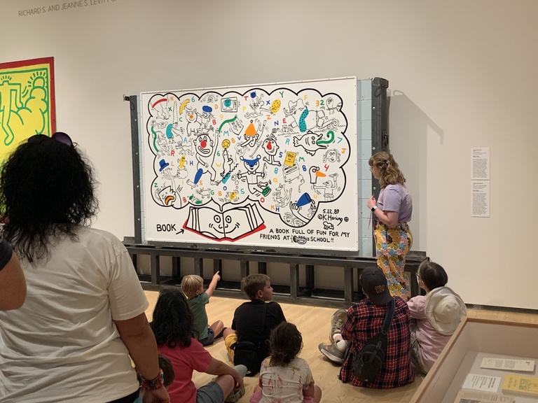 A photo of Bella Epstein, standing in front of the Keith Haring mural, "A Book Full of Fun," in the Stanley Museum of Art galleries, teaching in front of a group of children and their parents.