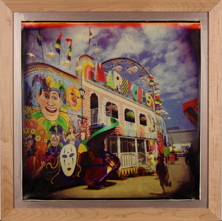 A two-story red brick building against a bright blue sky with fluffy white clouds. There is a colorful carnival themed mural painted on the front and a spiral slide from the top floor to the ground. A portion of a ferric wheel peeps out from behind the building.