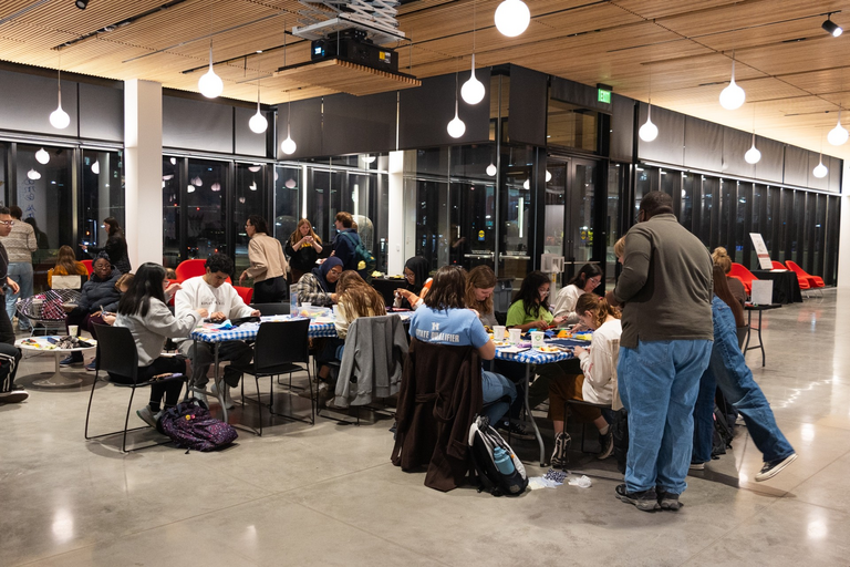 A view of the whole event; groups of students sit clustered around folding tables working on their projects; others mill about gathering supplies from the tables (fabric scraps, needles, thread) and chatting with other event attendees.