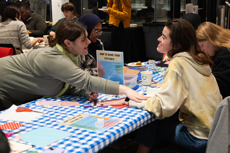 Two students, clearly friends, sit across from each other at a table at the event; one reaches across the table to hold the arms of the other excitedly.