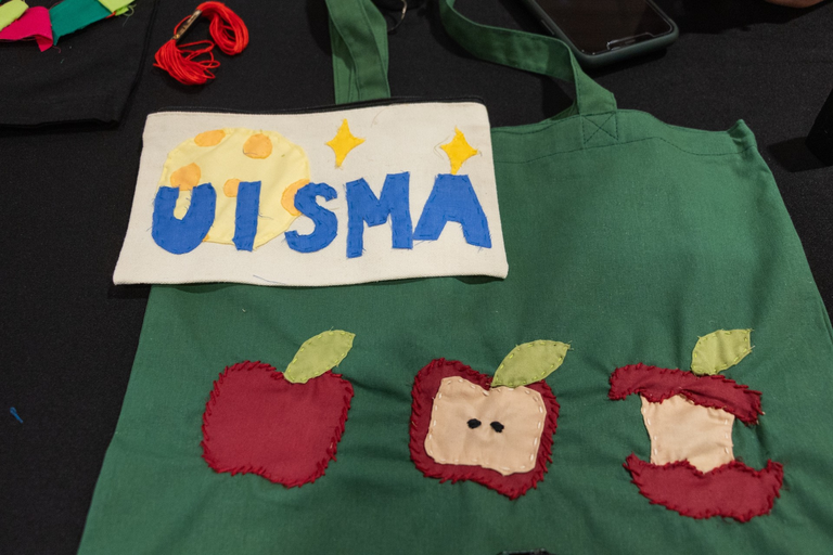 The example projects for the event: a white canvas pencil case with a moon and stars and blue text that spells "UI SMA" and a dark green tote bag with three apples, one whole, one halved, and one core, stitched onto it.