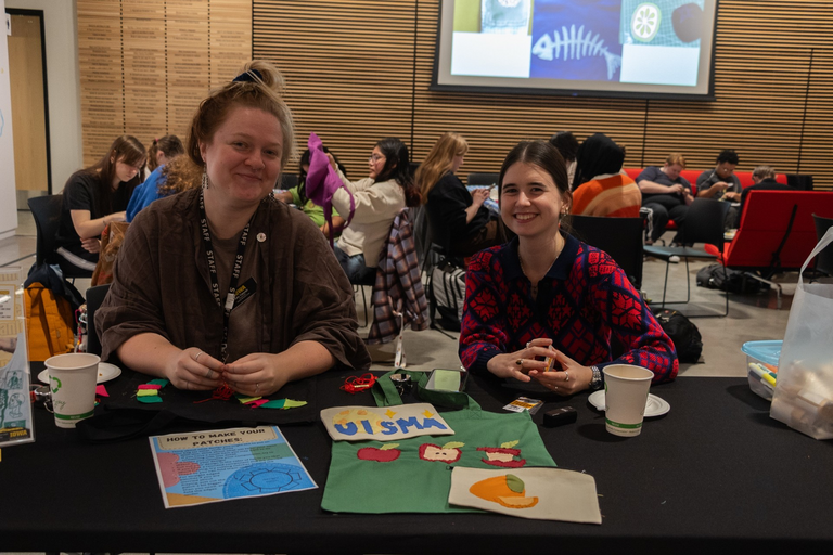 A view of the event from the welcome table: Campus Engagement Coordinator Annelies Knight and Assistant Curator of Student Engagement Allie Tokarski sit ready to welcome students to the event that is happening behind them.