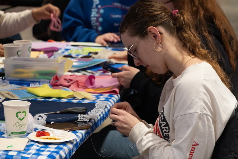 A photo of a student, in profile, working on their sewing project; fabrics, sewing supplies, and other materials are scattered in front of them on a table covered with a blue gingham tablecloth.