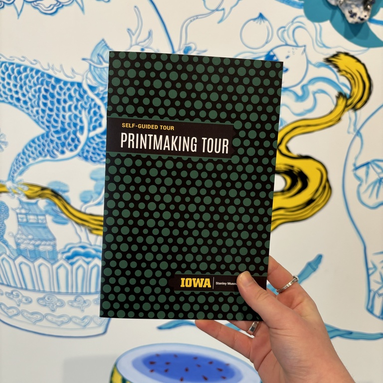 A hand holds a copy of the printmaking tour in front of the lobby mural at the Stanley.
