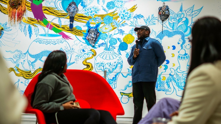 Person in a denim jacket speaks to a seated audience in front of a colorful wall mural with floral and abstract designs.