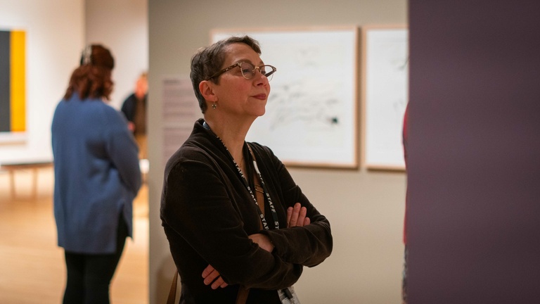 A person in a black dress with glasses observes an artwork on wall in an art museum