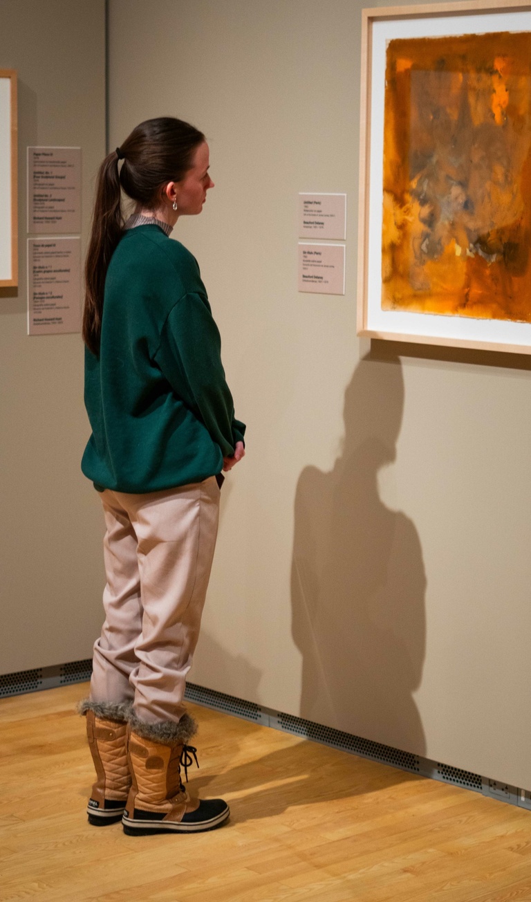 A person wearing a green sweater and beige pants observes a painting on a wall in an art museum