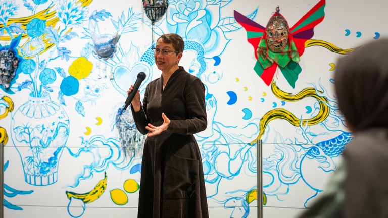 Person in a black dress speaks to an audience in front of a colorful wall mural with floral and abstract designs.