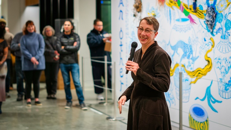 Person in a black dress holding a mic speaks to an audience in front of a colorful wall mural with floral and abstract designs.