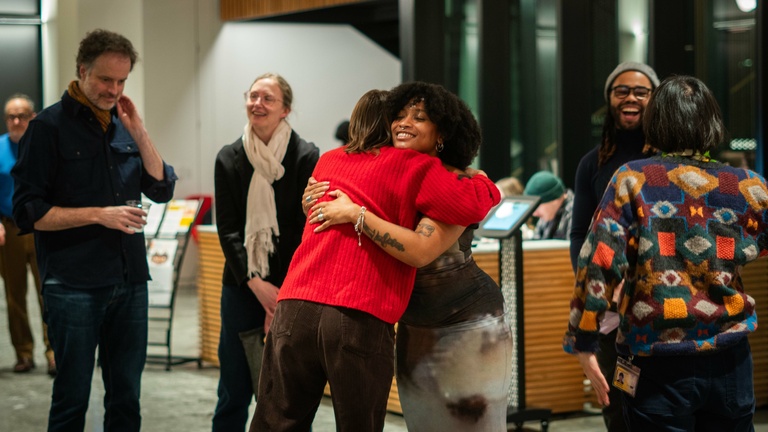 A person in a red sweater embraces a woman in a dress as people stand around them smiling and chatting