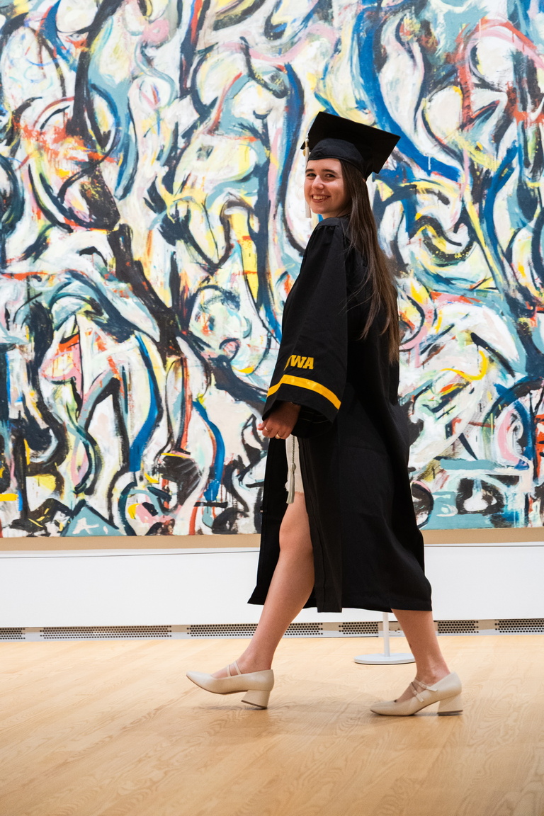 Gallery Host and Campus Engagement Coordinator Annelies Knight poses in her graduation gown in front of Jackson Pollock's Mural.