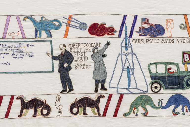 Embroidered tapestry panel depicting two men in a blue suit and grey coat. One points to a blackboard on the left with mathematical equations scribbled on it, and the other points to an early model of a rocket hoisted on a frame structure. alongside early cars, dinosaurs, and the text "ROBERT GODDARD BUILT THE FIRST LIQUID FUEL ROCKET."