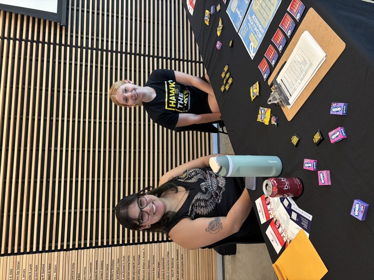 Two members of student group Hawk The Vote sit at a table, smiling.