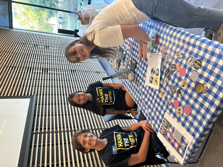 Two members of Hawk the Vote assist an event attendee with using a pinback button press.