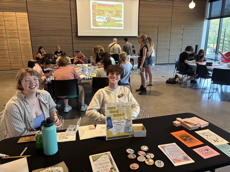 An overview of the event: two students sit at a welcome table, while various others mill about behind them working on crafts.
