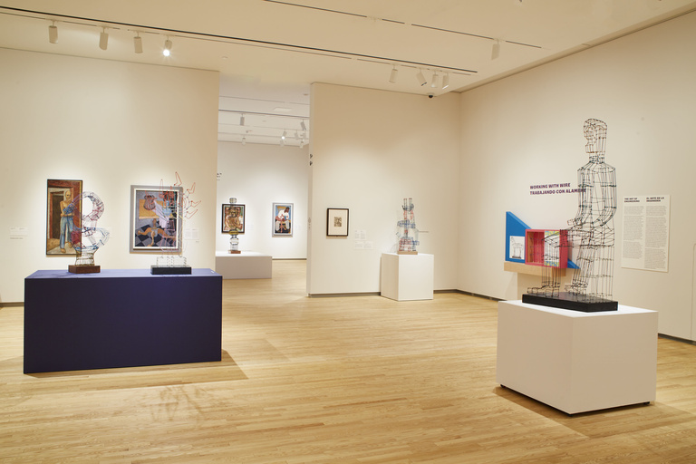 An open gallery space with light wooden floors and colorful paintings on white walls. There are geometric wire sculptures displayed on blue and white pedestals towards the center of the room.