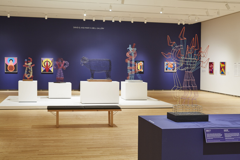 An open gallery space with light wooden floors and colorful abstract paintings on blue and white walls. There are geometric wire sculptures displayed on white pedestals and a blue platform towards the center of the room.