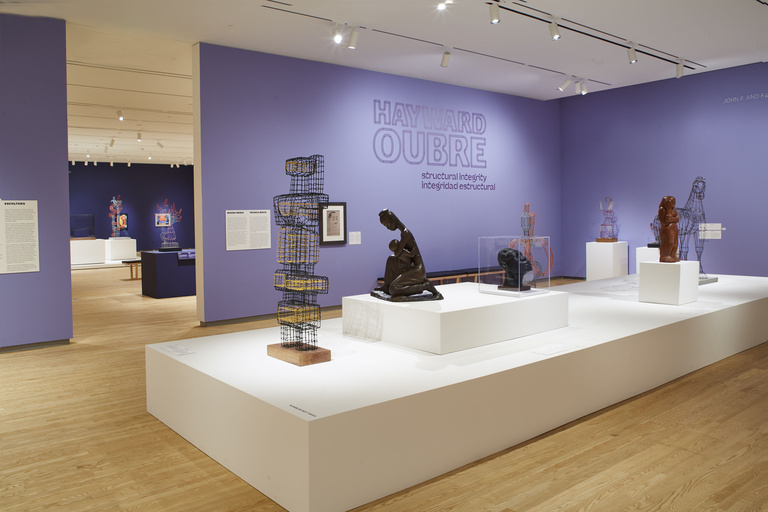 An open gallery space with light wooden floors displays wire and metal sculptures on white platform in the center of the room, with purple accent walls and the text "Hayward Oubre: Structural Integrity" featured prominently on the wall.