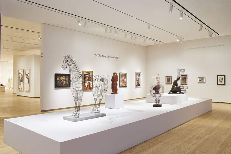 An open gallery space with light wooden floors and white walls displays wire and metal sculptures on a large white platform in the center of the room, with the text "Art is long, life is short" featured prominently on the back wall.