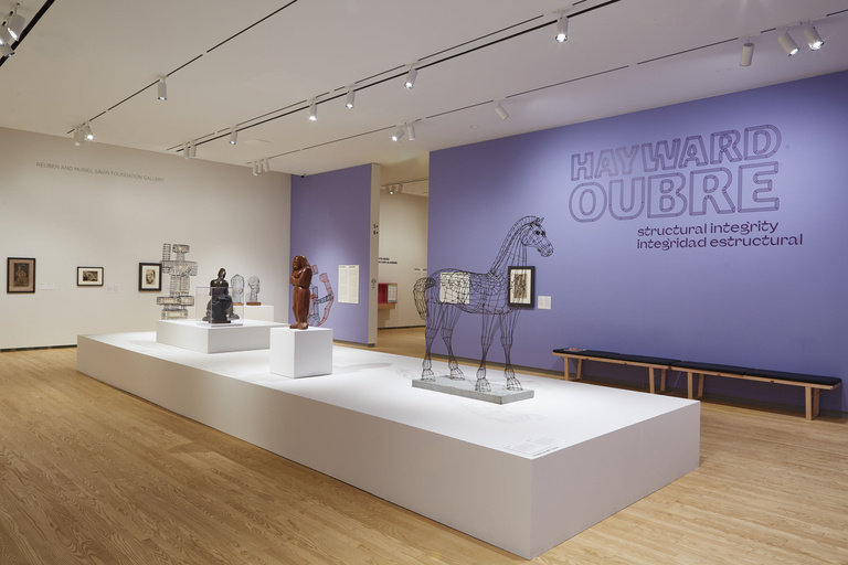 An open gallery space with light wooden floors displays wire and metal sculptures on white platform in the center of the room, with purple accent walls and the text "Hayward Oubre: Structural Integrity" featured prominently on the wall.