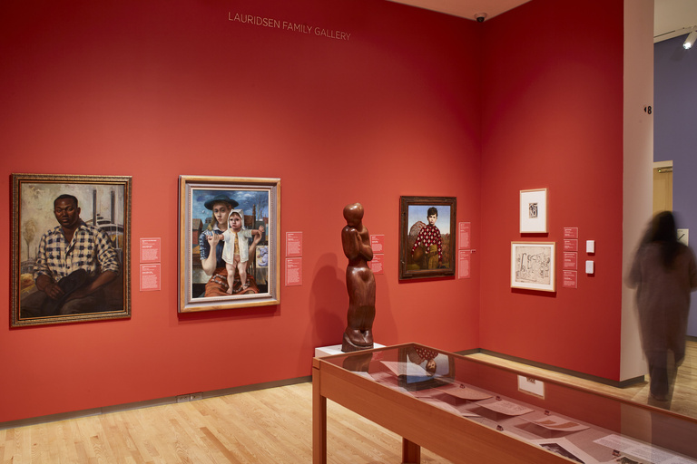 An art gallery space with light wooden floors featuring paintings, a sculpture, and display case against red walls.