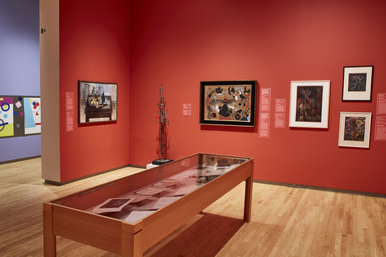 An art gallery space with light wooden floors featuring paintings, a sculpture, and display case against red walls.