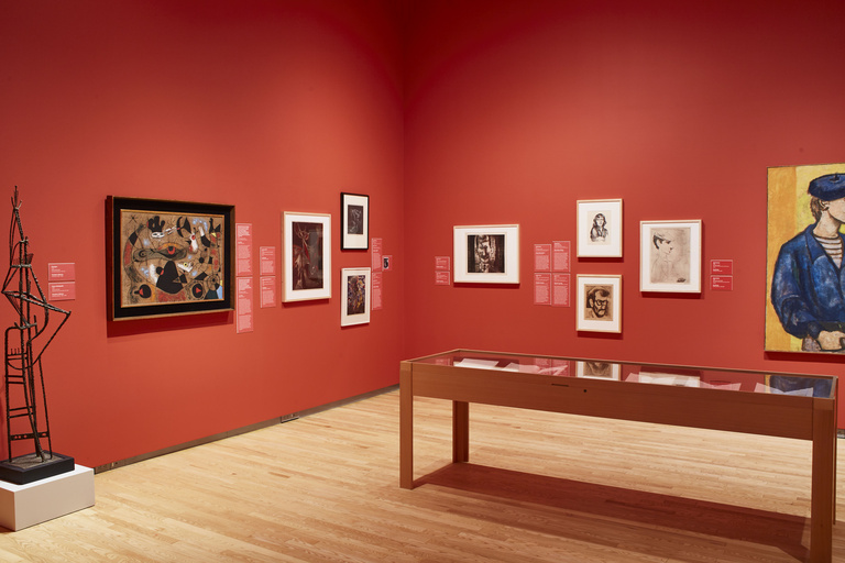 An art gallery space with light wooden floors featuring paintings, a sculpture, and display case against red walls.
