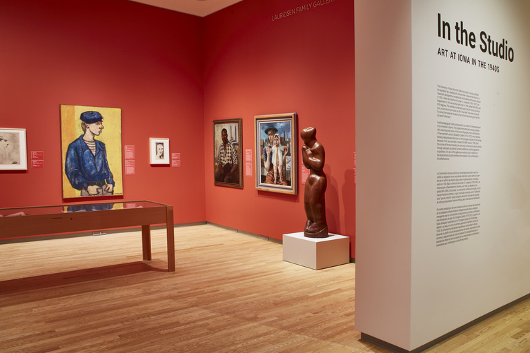 An art gallery space with light wooden floors featuring paintings, a sculpture, and display case against red walls. A white baffel wall encompasses the exhibition space, with vinyl text featured on it that reads "In the Studio: Art at Iowa in the 1940's."