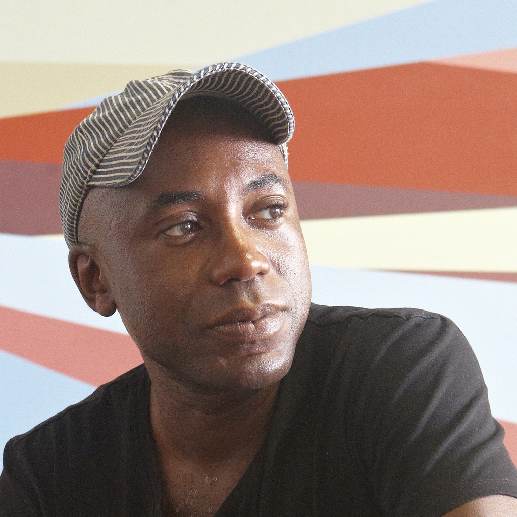 A Black man in a dark t-shirt and beige ball cap in front of a mural painted with geometric shapes in bright colors.
