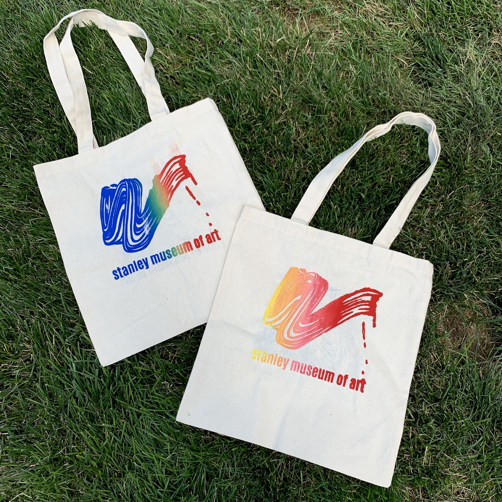 A photograph of two Stanley tote bags, hand printed by the Stanley Campus Council, laid out on bright green grass. One is printed with a rainbow ink gradient, and the other with a gradient of reds and yellows. Both read "Stanley Museum of Art" beneath a stylized, drippy brushstroke motif.