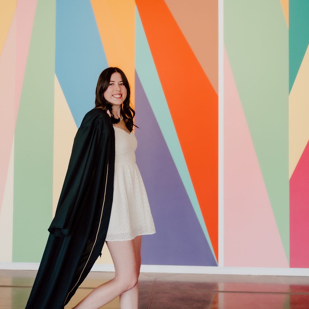 A photo of Anaka Sanders; it is an action shot of her striding in front of the lobby mural at the Stanley, dressed in her graduation robes.