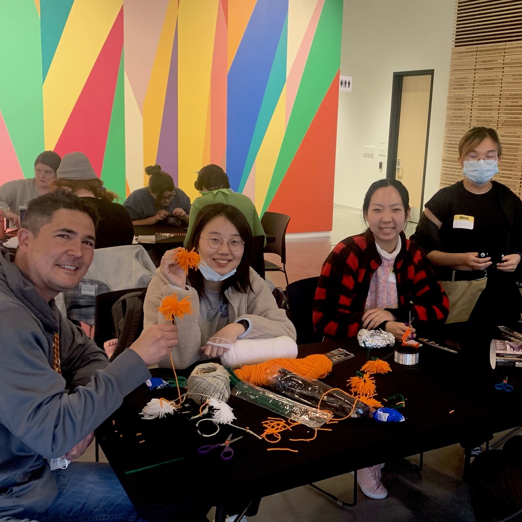 A photo of four students gathered around a folding table in the Stanley Museum of Art lobby. They are all in the midst of making crafts: cutting yarn to make pompoms and assembling Halloween garlands.