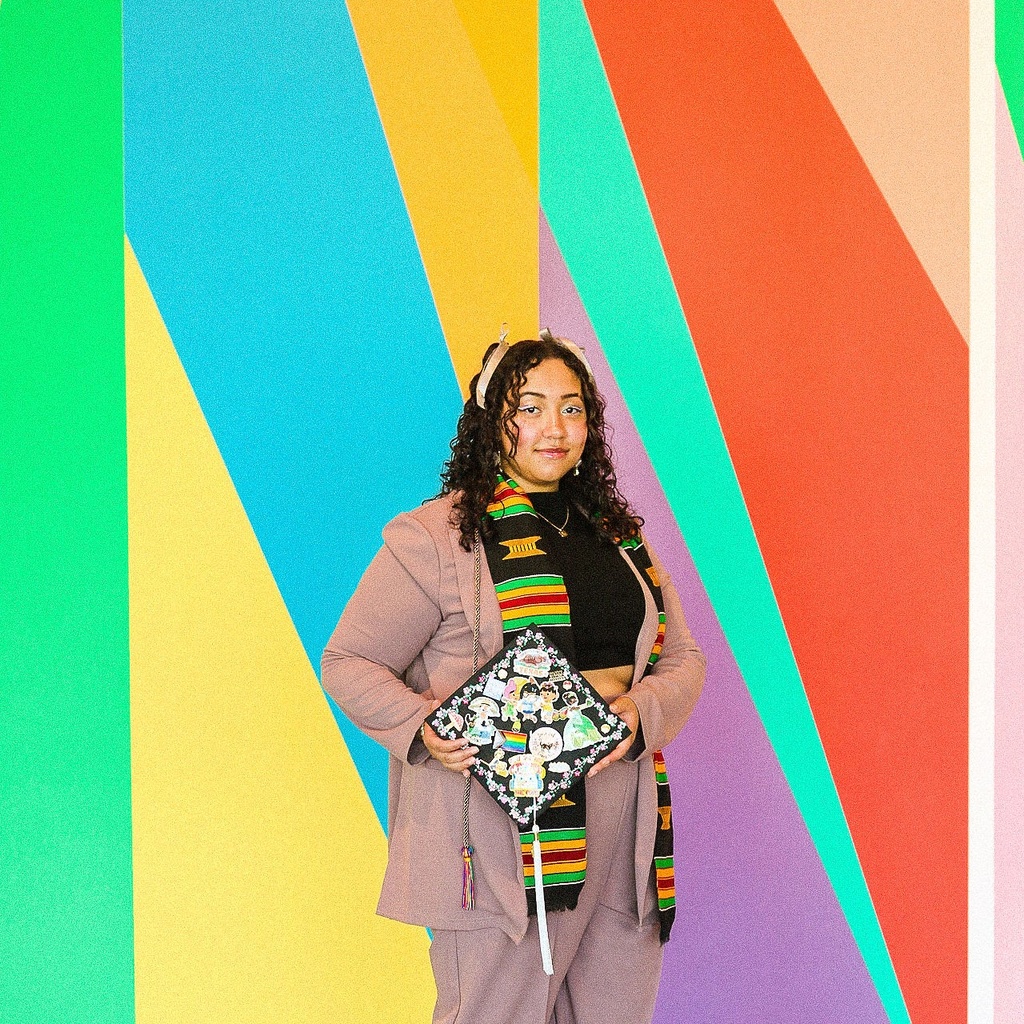 A photo of Lily Hester: they are standing in front of the mural in the Stanley's lobby, dressed up, posing with their graduation cap.