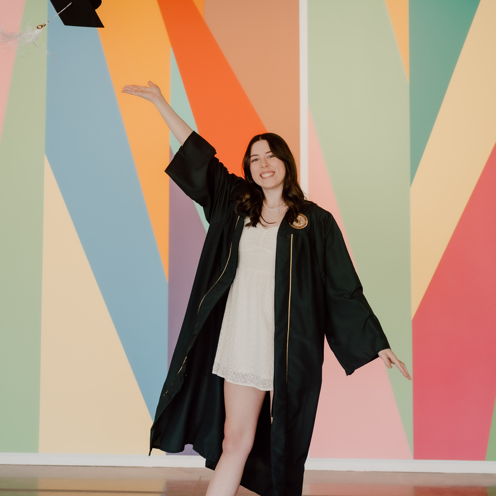 A photo of Anaka Sanders; she is posing in front of the lobby mural at the Stanley, dressed in her graduation robes, tossing her cap in the air.