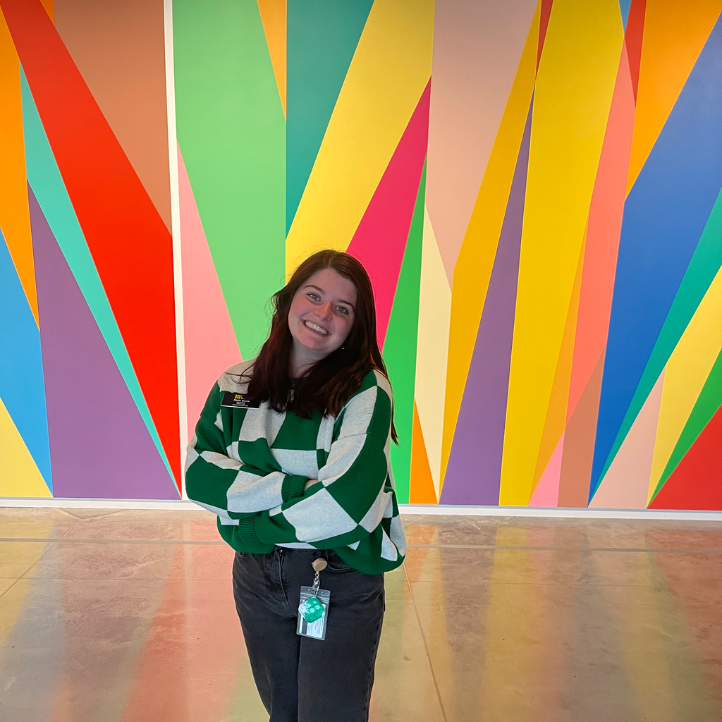 A photo of Alexis Belme: she is standing in front of the mural in the Stanley Museum of Art lobby, smiling, her arms crossed.