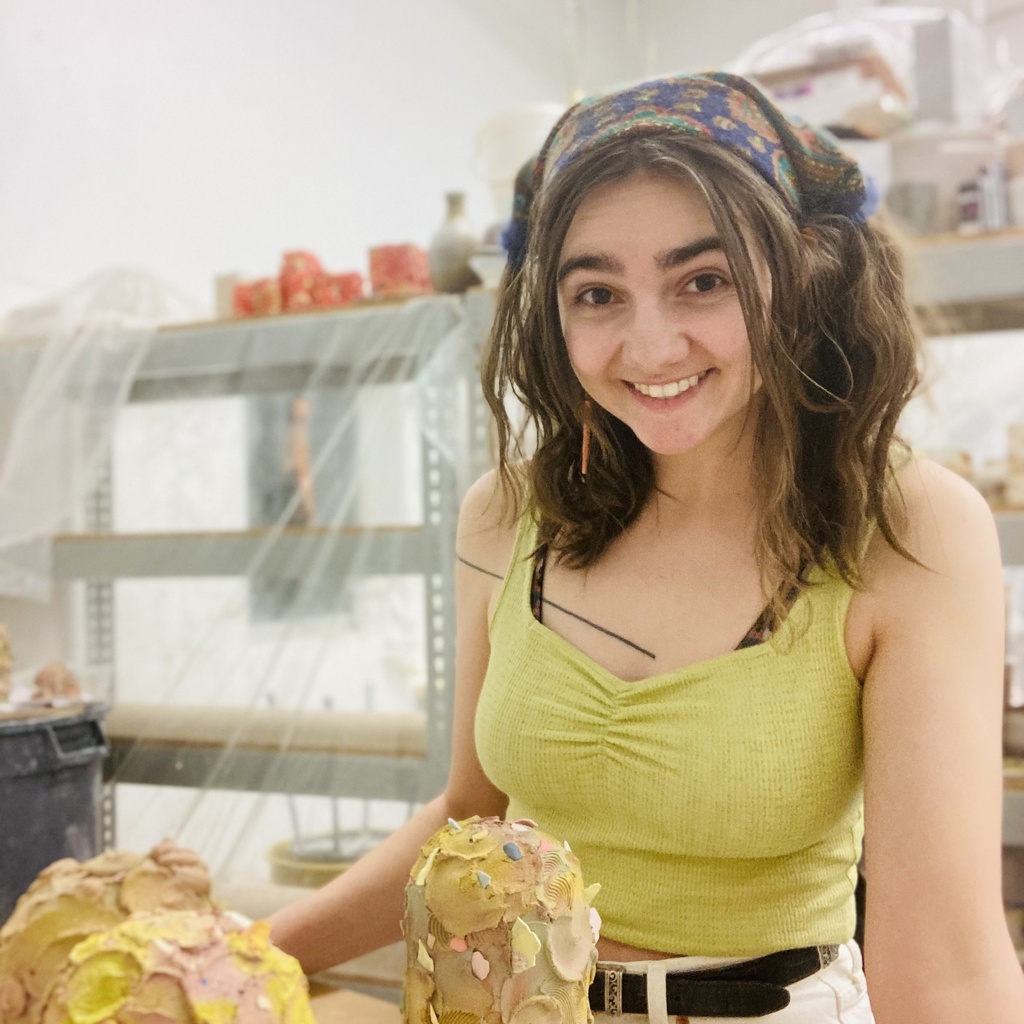 A photo of Alyson Holevoet: she poses in a ceramics classroom with some pieces she is working on.
