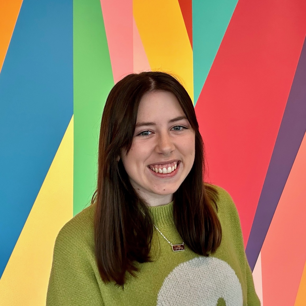A photo of Anaka Sanders: she is wearing a green knit sweater with a smiley face on the front and standing in front of the colorful mural in the lobby of the Stanley Museum of Art.