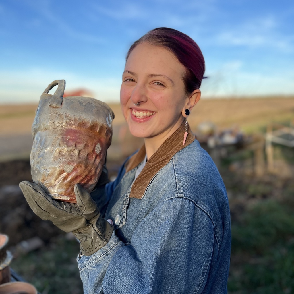 A photo of Eliza Link: she is smiling at the camera, holding a large pot with oven mitts.