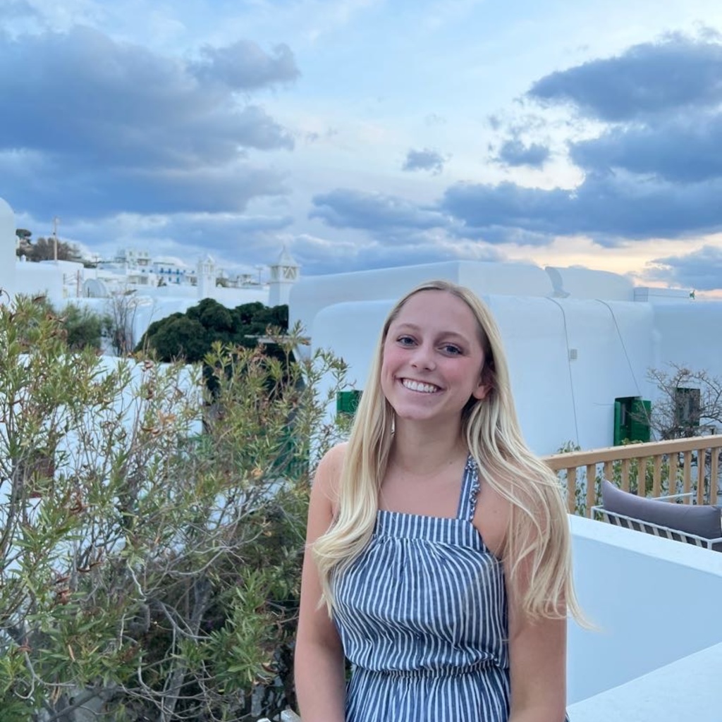 A photo of Jenna Mosnik: she is wearing a blue dress and smiling, posing in front of a beautiful body of water.