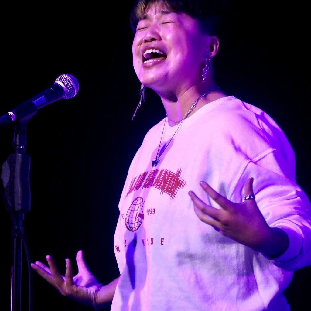 A photo of Samm Yu: bathed in pink light, they are standing in front of a microphone, speaking and gesticulating.