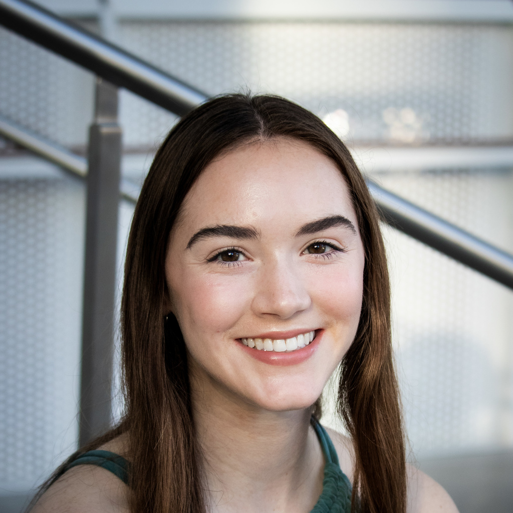 A photo of Katherine Shamdin; she is seated on stairs, smiling at the camera.