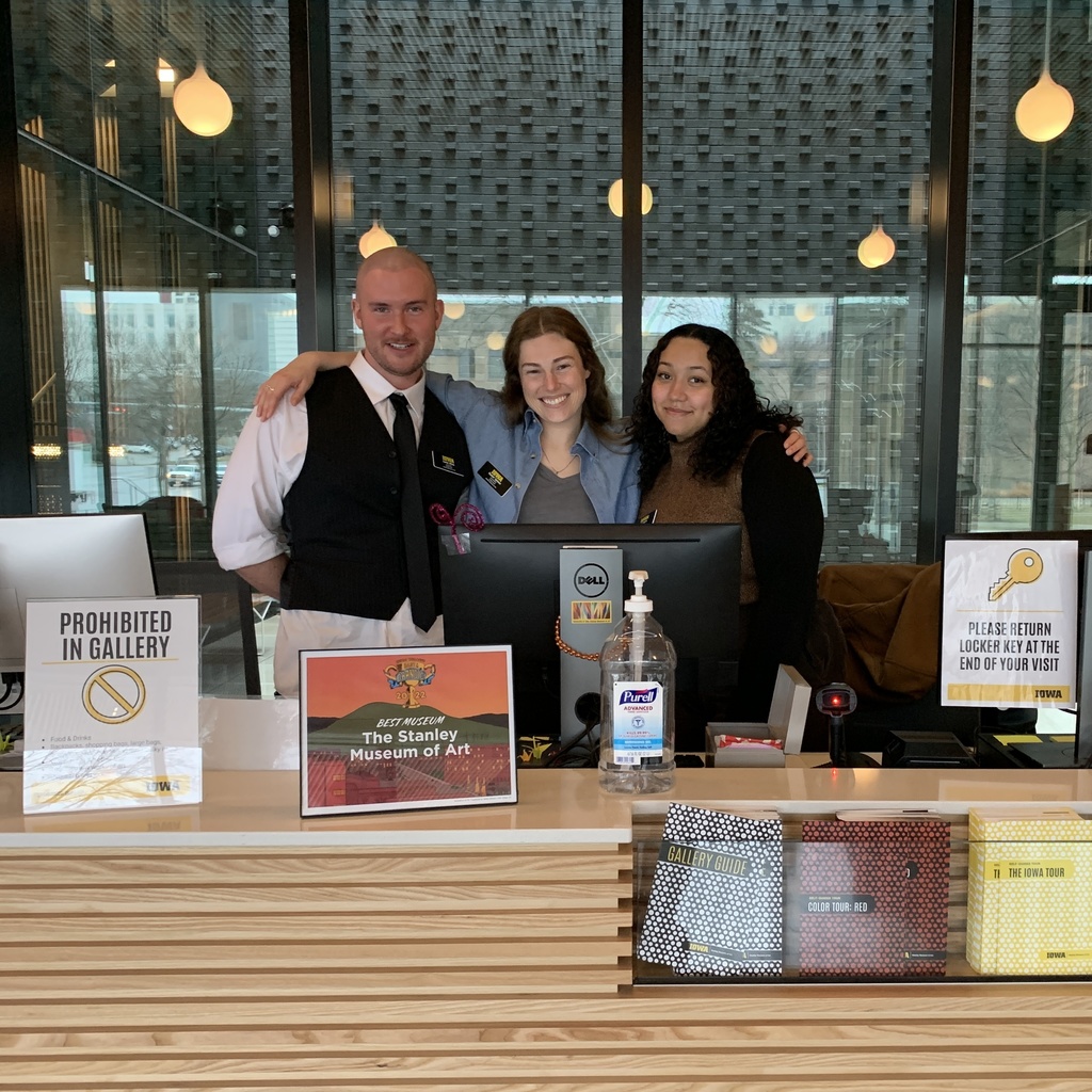 A photo of three gallery hosts standing at the Stanley Museum of Art front desk. Carter, Rachel, and Lily are crowded together, Rachel's arms around Carter's shoulders on her right, and Lily's shoulders on her left.