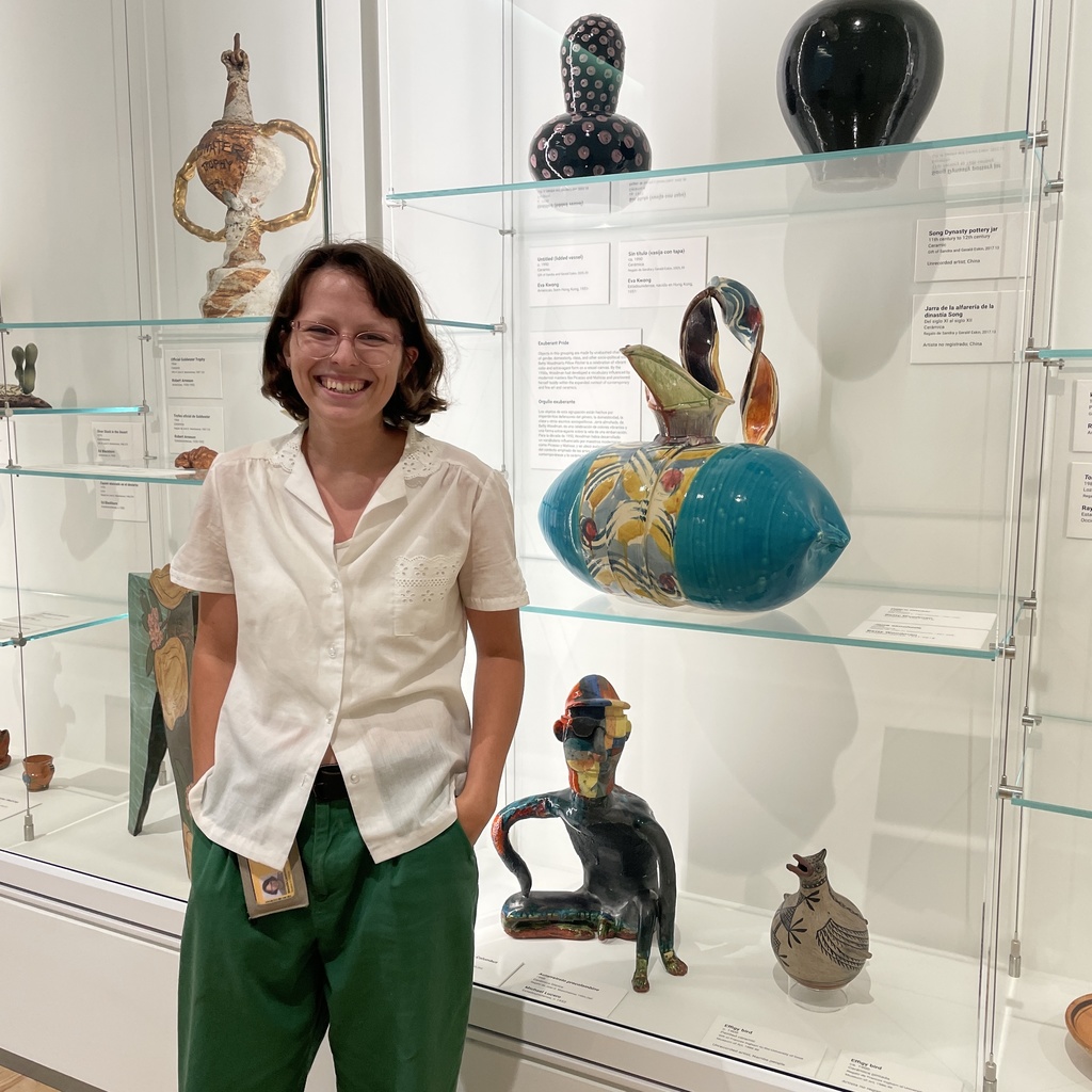 a photo of Melanie, smiling and posing in front of a glass display case of ceramics.
