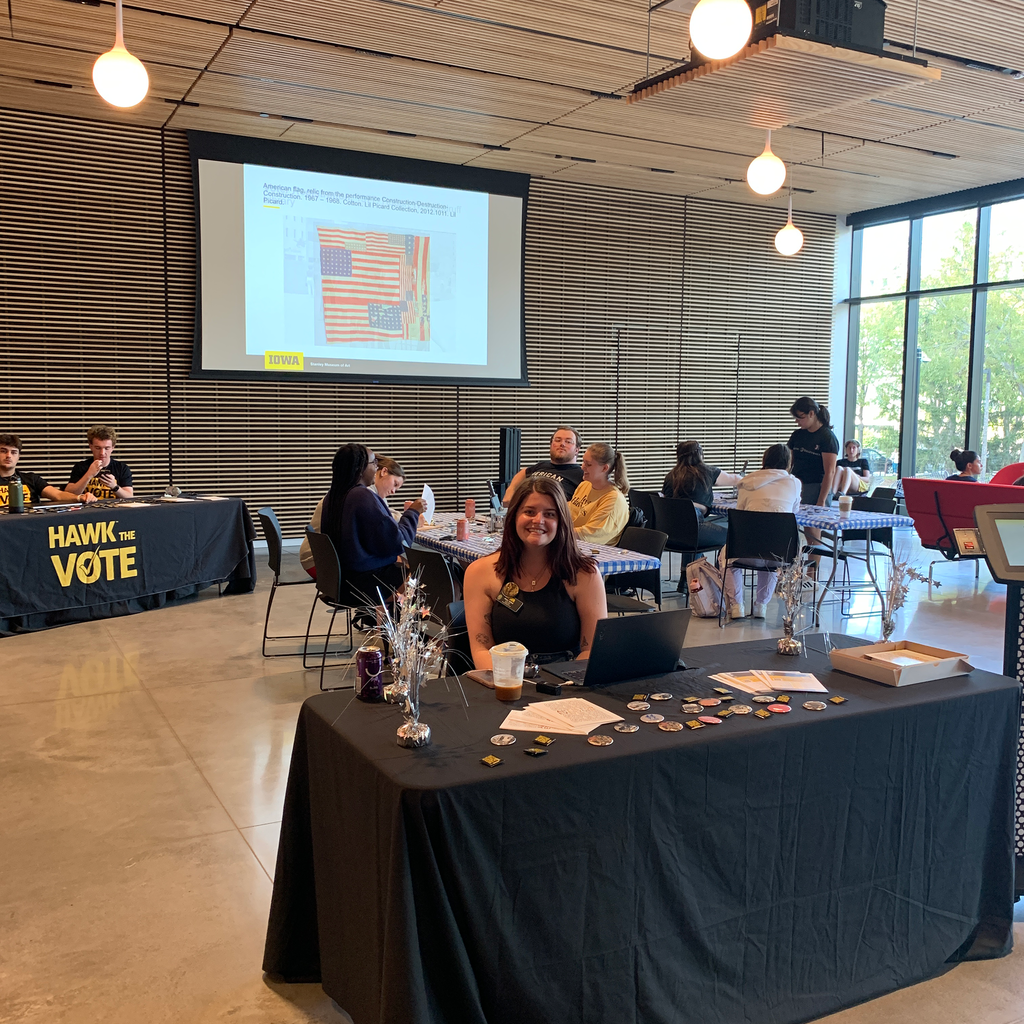 A photo of the Art of Voting event, showcasing the info table at the front of the event. At the table sits Campus Engage Coordinator Alexis Belme, who is smiling, prepared to greet event attendees as they walk through the museum's front doors.