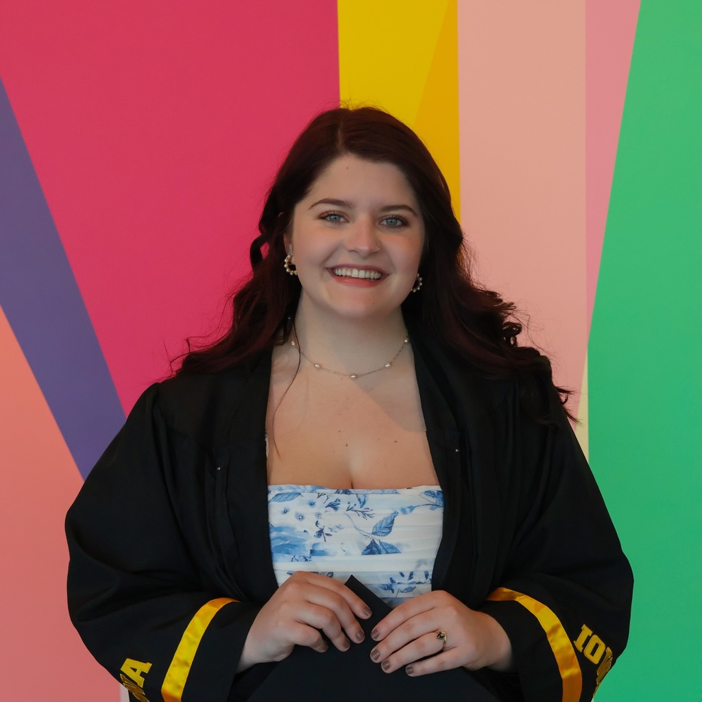 A photo of Alexis Belme in her graduation cap and gown; she poses, smiling, in front of the multi-colored mural in the Stanley Museum of Art lobby.