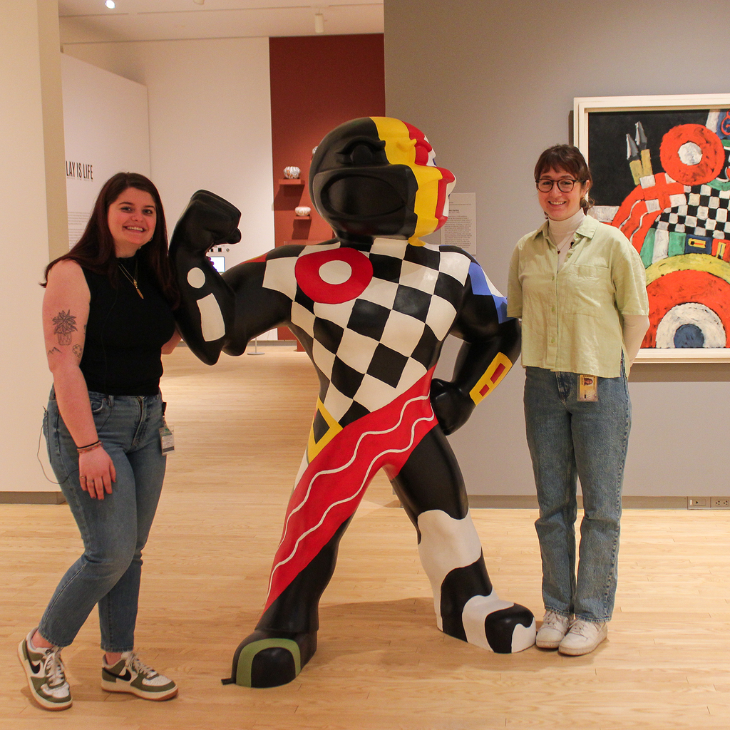 Two students stand in the galleries at the Stanley Museum of Art, posing next to a 6-foot-tall fiberglass Herky statue, that is painted to match a painting in the background of the photo--Marsden Hartley's "E."