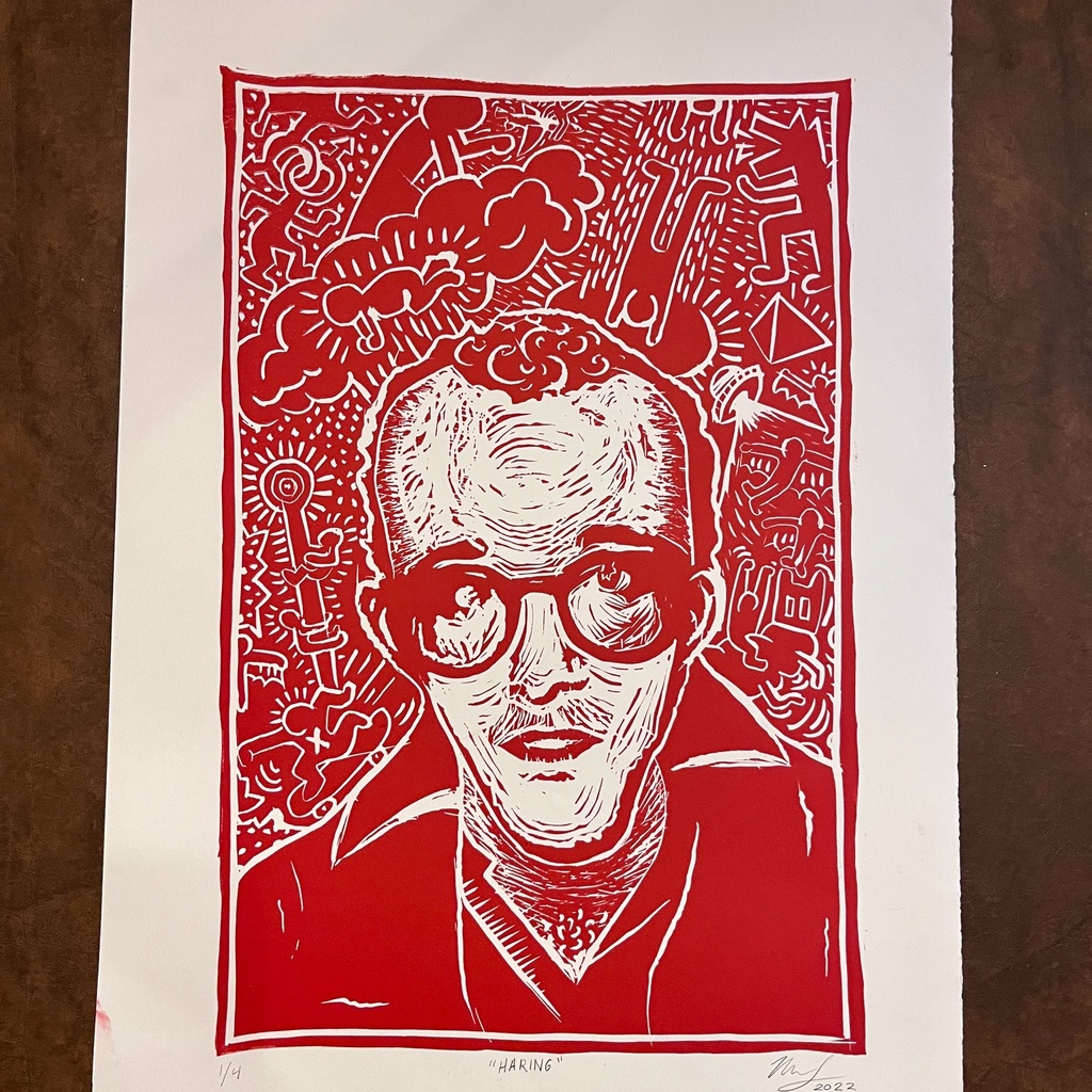 A photo of a linoleum block print; printed in red ink, the image is of Keith Haring, a young man wearing glasses, with vibrant, childlike imagery, reminsicent of his artwork, swirling around his head. 