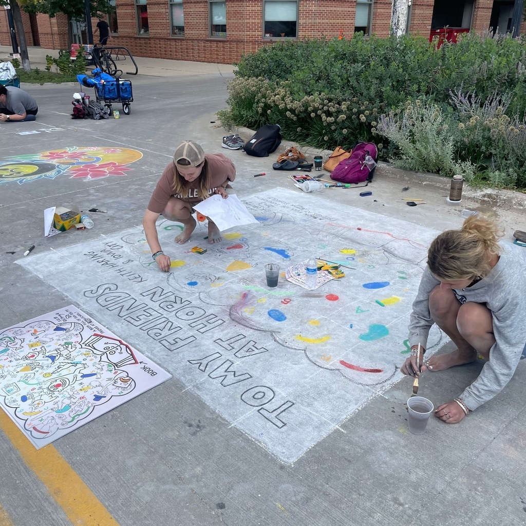 Student employees Sarena Gibson and Mari Johnson crouch on the street in downtown Iowa City, where they are working on a large chalk drawing as part of the city's Rock the Chalk summer festival.