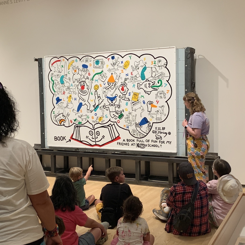 A photo of Bella Epstein, standing in front of the Keith Haring mural, "A Book Full of Fun," in the Stanley Museum of Art galleries, teaching in front of a group of children and their parents.