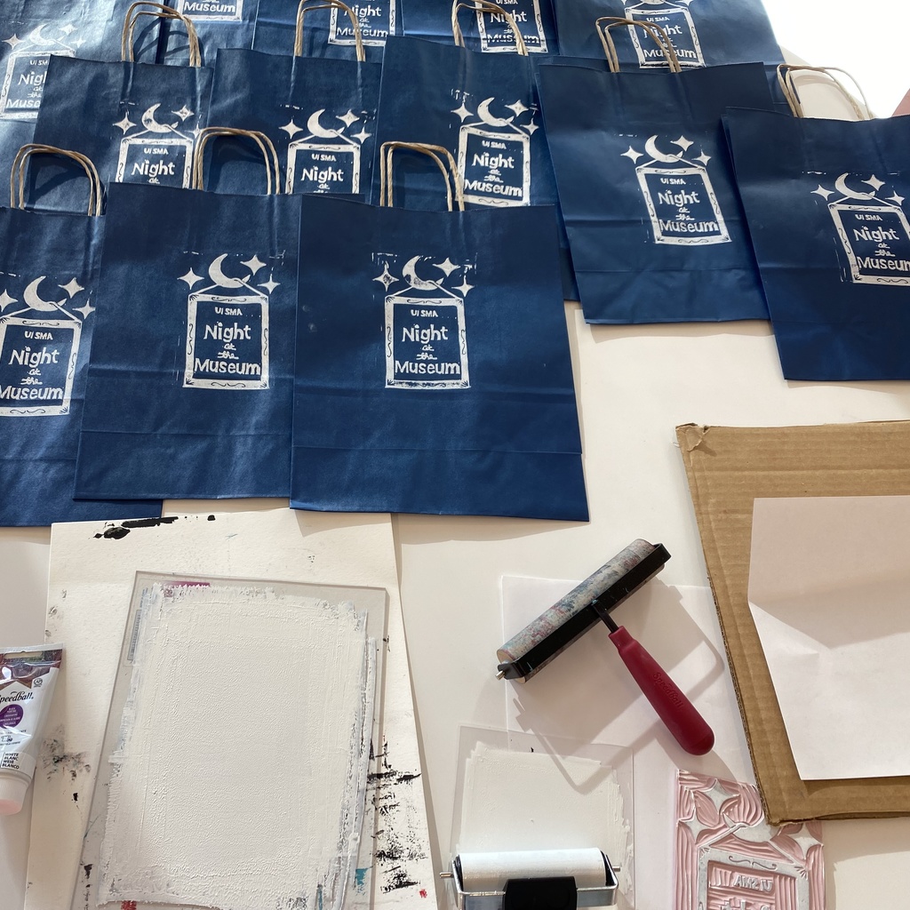 A photo of the entire setup: approximately one dozen printed blue paper bags are lined up along the top of a table, waiting to dry, set above a bottle of white ink, a plate with white ink on it, the ink covered linoleum block, an inked up brayer, and a clean brayer that has been used to apply pressure to the back of the plate to help transfer the ink.